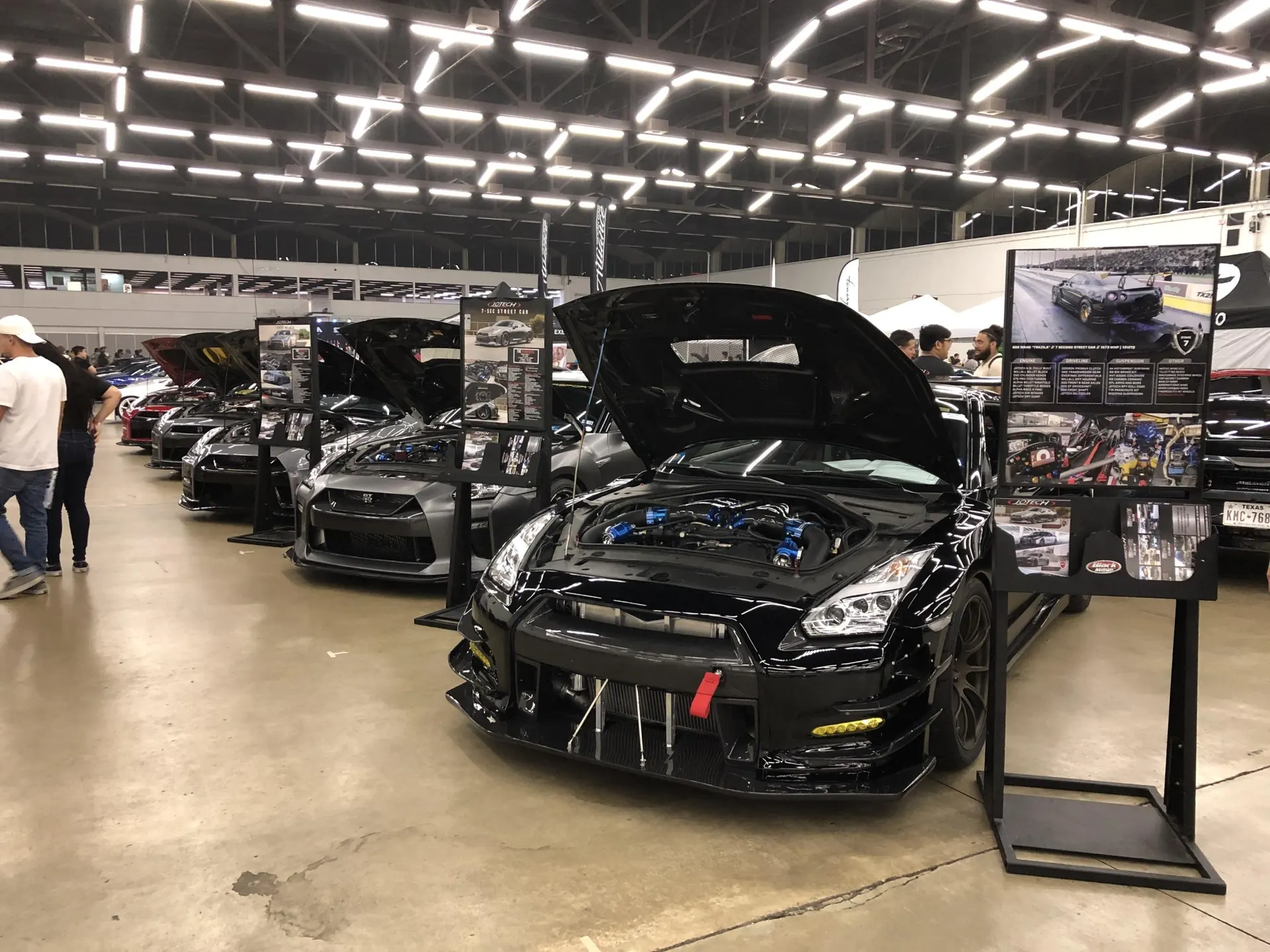 Modified Nissan GT-R with an open hood showcasing a custom turbo and boost piping setup at an indoor car show.