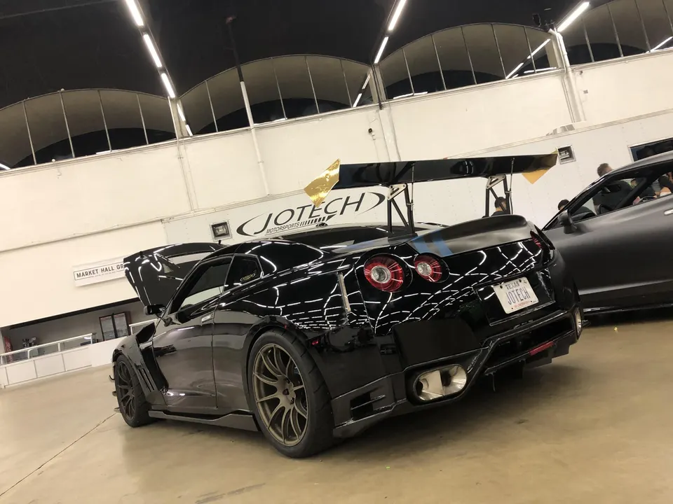 Black Nissan GT-R with widebody kit, large aftermarket wing, and custom wheels displayed at a Jotech Motorsports event.