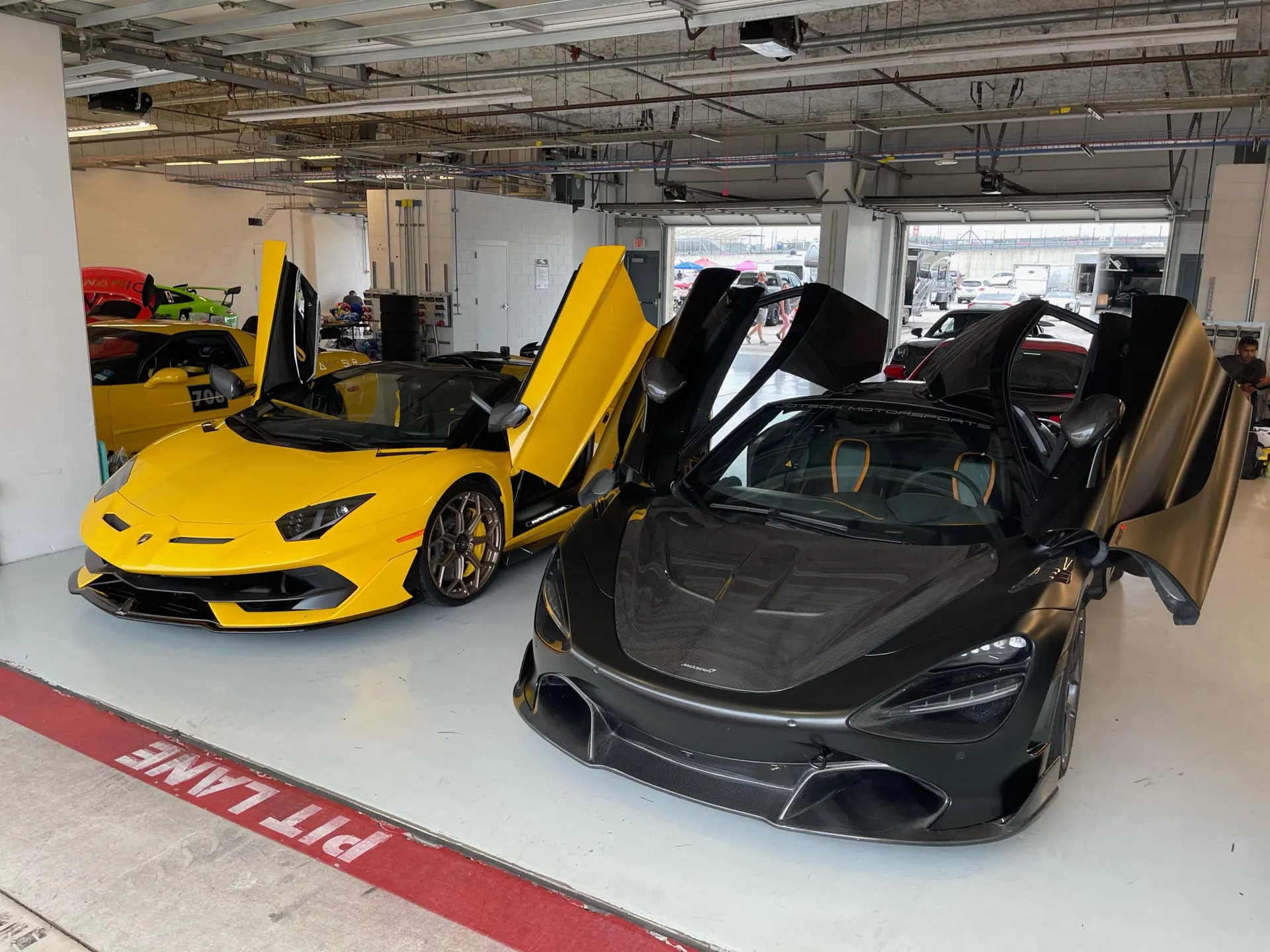 Yellow Lamborghini Aventador and black McLaren 720S with open doors parked in a pit lane garage at a track.