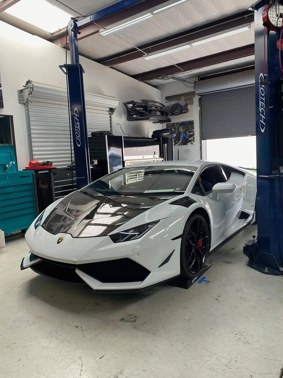 A white Lamborghini Huracan with a carbon fiber hood and front aero kit parked in a Jotech Motorsports shop.