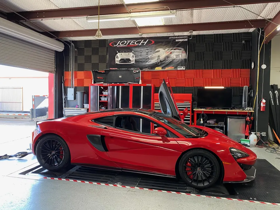 A red McLaren supercar with its doors open, positioned on a dyno inside the Jotech Motorsports performance tuning shop.