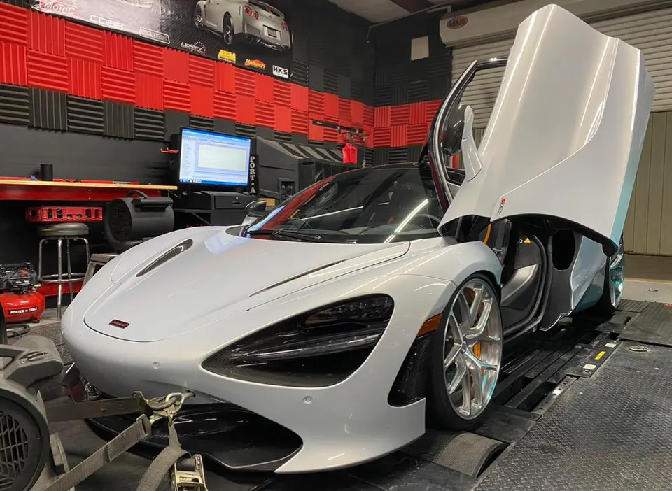 A silver McLaren 720S with its butterfly doors open, strapped to a dynamometer for performance tuning in a shop.