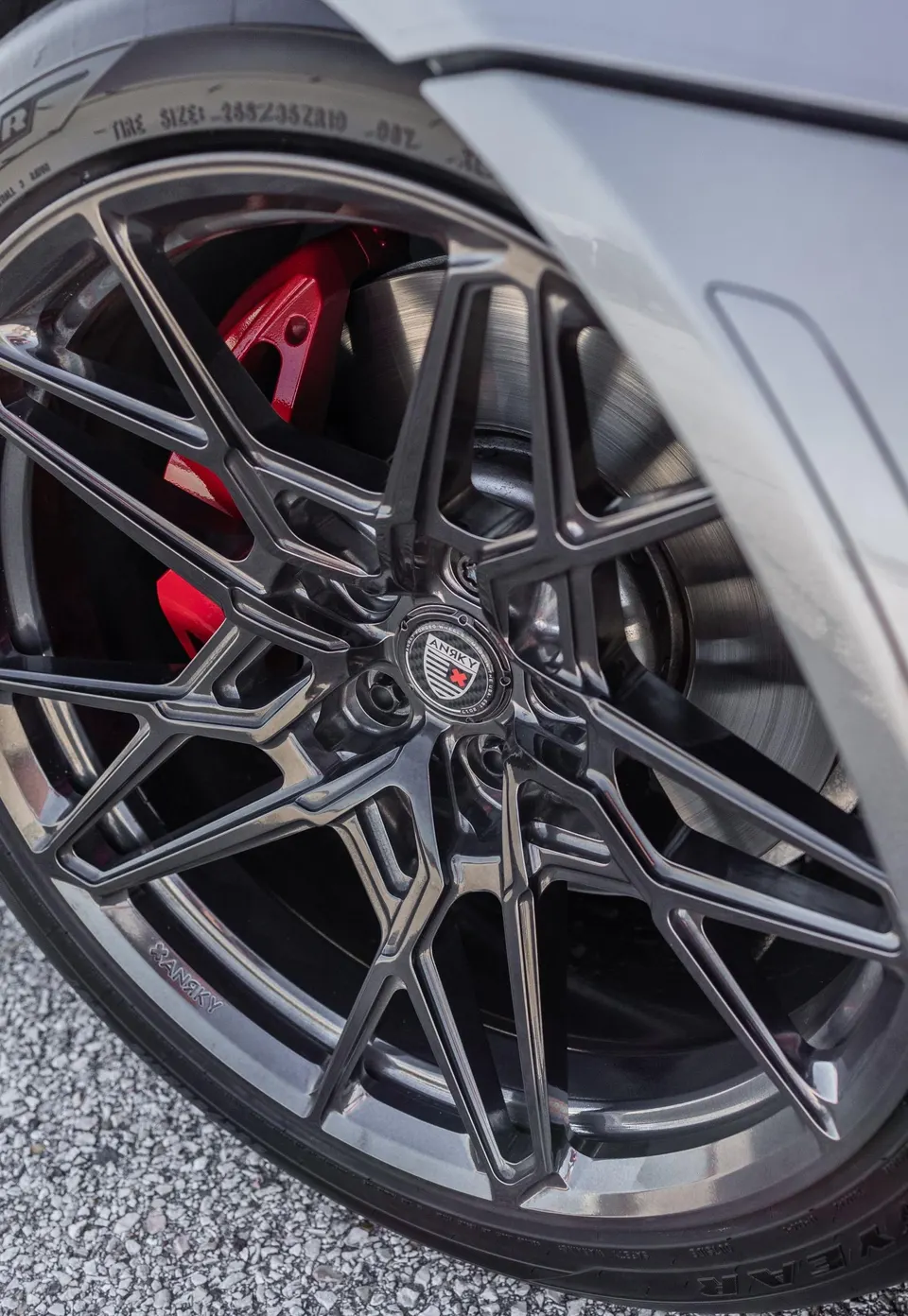 A close-up of a custom ANRKY wheel with red brake calipers and a performance tire on a grey car.