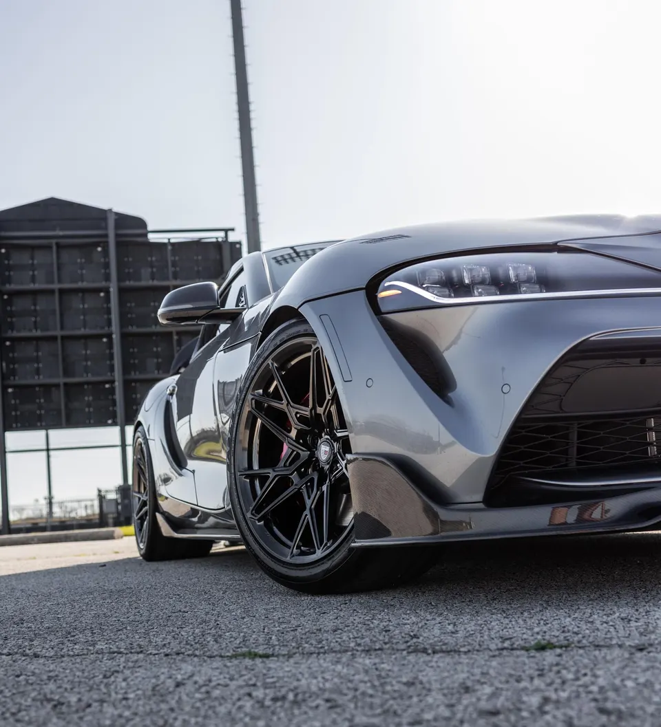 Grey Toyota Supra A90 with aftermarket black multi-spoke wheels and carbon fiber front splitter parked outdoors.
