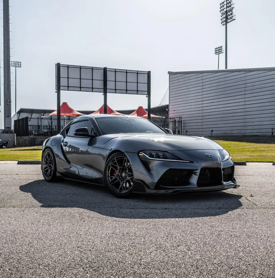Grey Toyota Supra MK5 with aftermarket wheels and front splitter parked outdoors near a stadium.