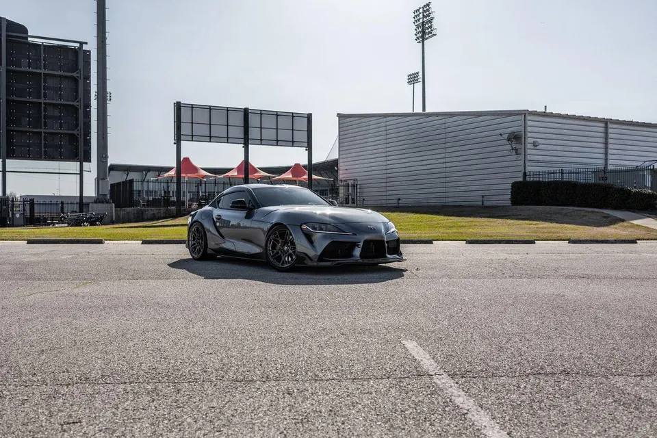Grey Toyota Supra MK5 parked in a lot with stadium lights and buildings in the background, showcasing its sleek design.