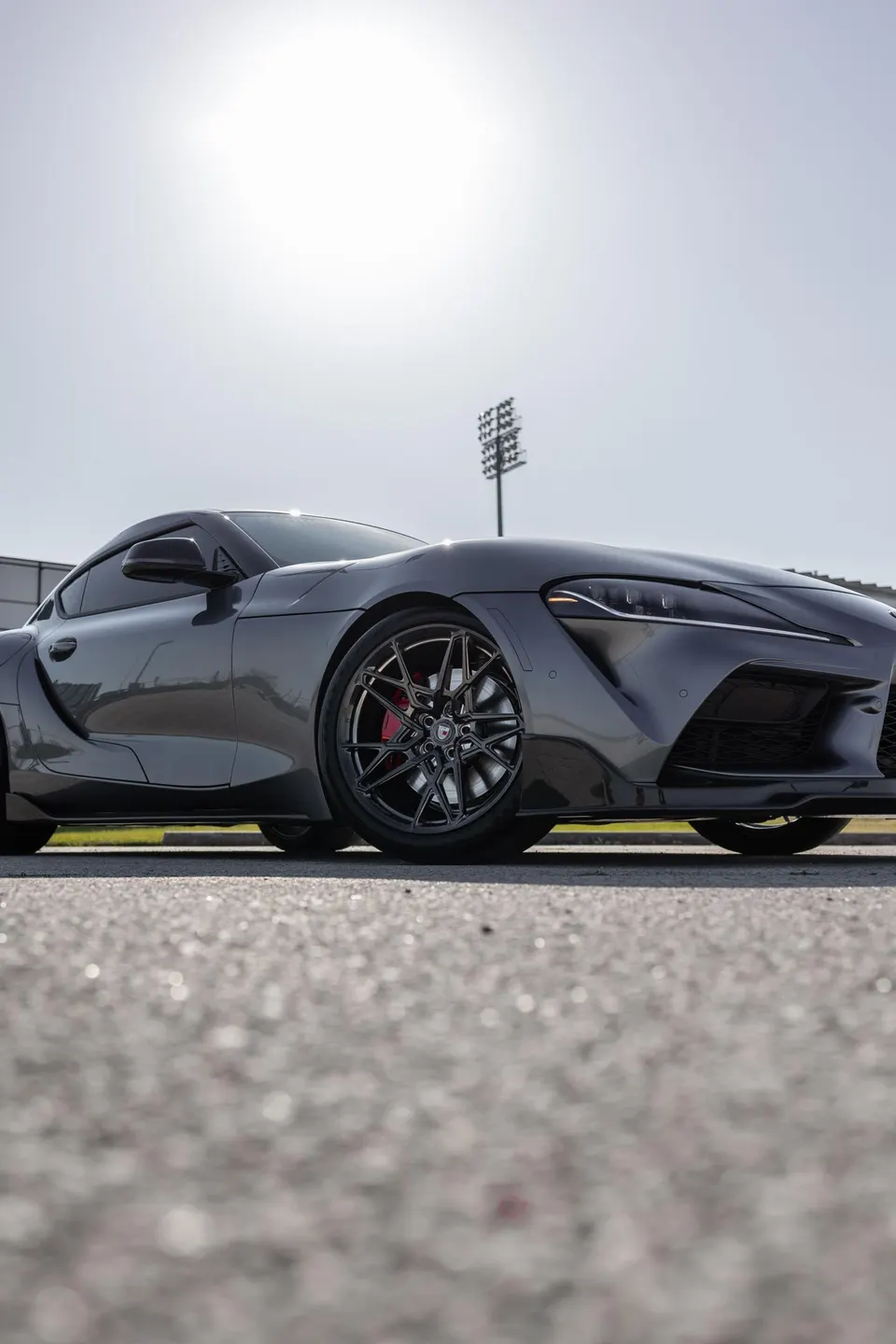 Dark grey Toyota Supra with aftermarket multi-spoke wheels and red brake calipers parked outdoors under bright sunlight.