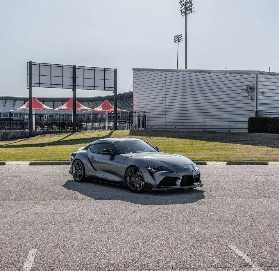 A grey Toyota Supra with aftermarket wheels parked in a lot outside a stadium on a sunny day.