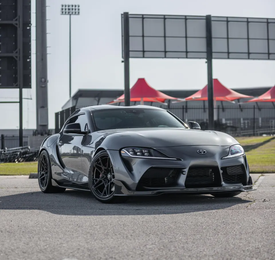 A grey Toyota Supra with aftermarket wheels and a front splitter parked at a track venue on a sunny day.