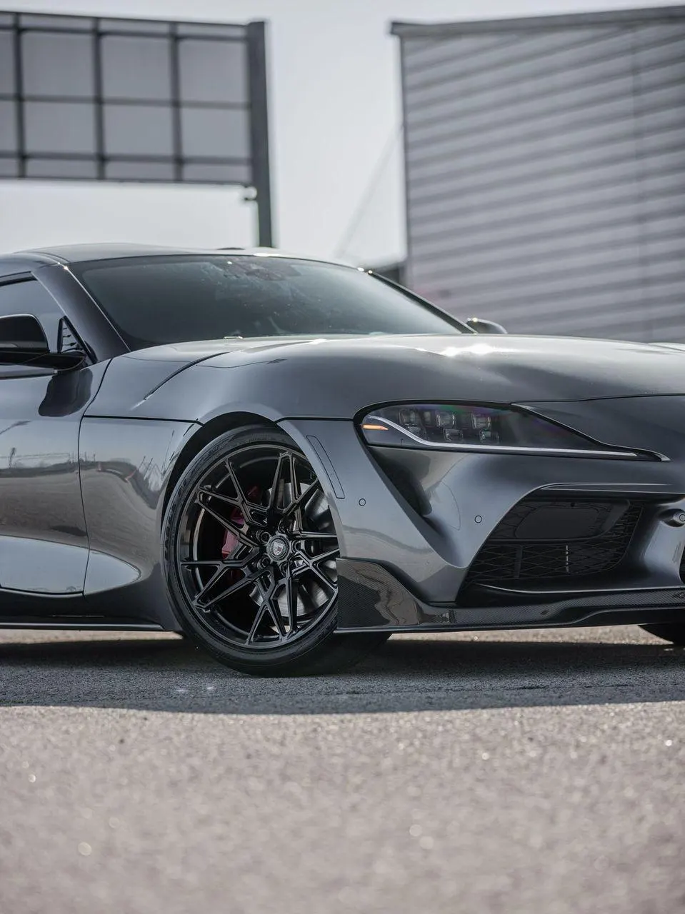 Close-up of a grey Toyota Supra A90 featuring aftermarket black multi-spoke wheels, red brake calipers, and a carbon fiber front lip.