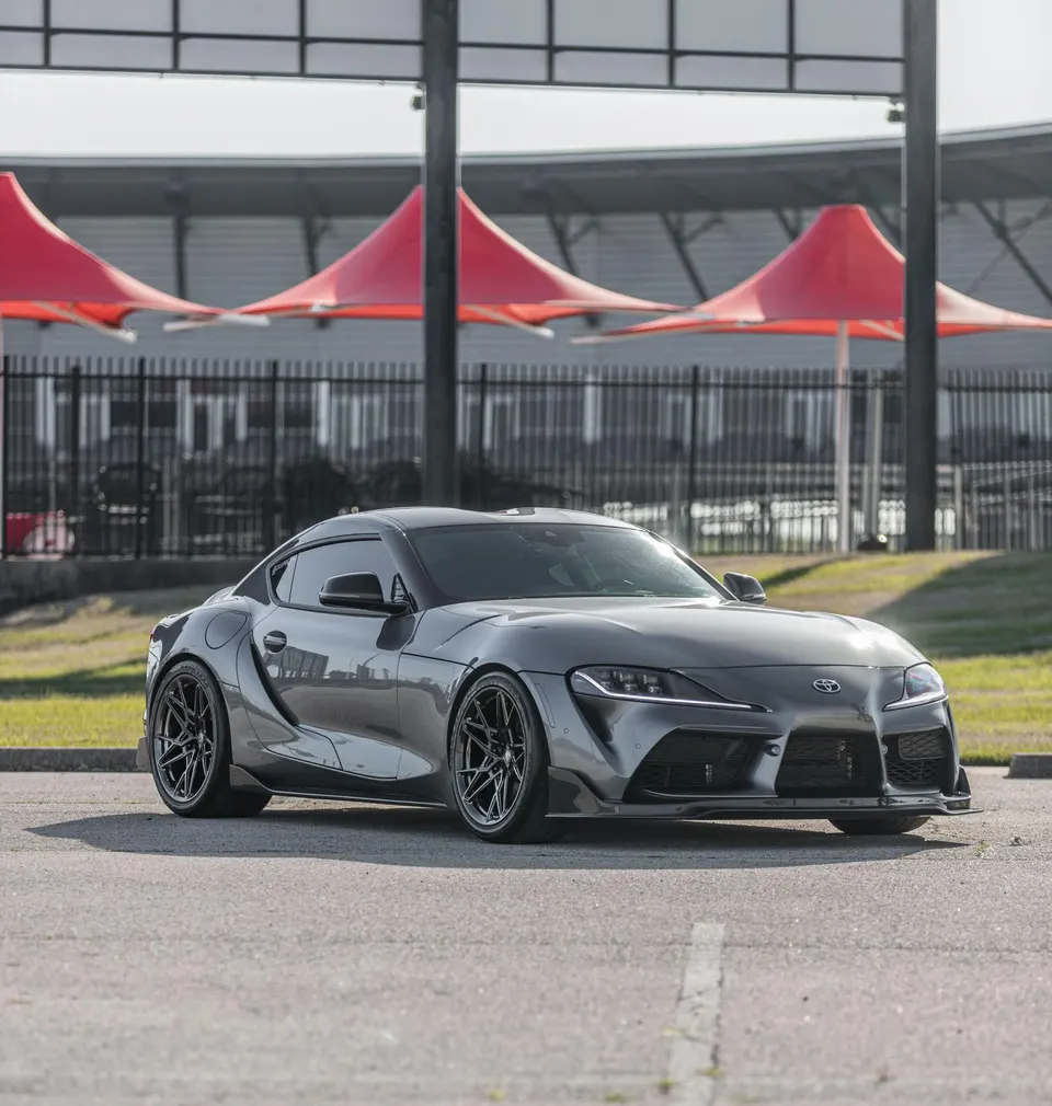 Grey Toyota Supra with aftermarket wheels and a front splitter parked outdoors at a venue.