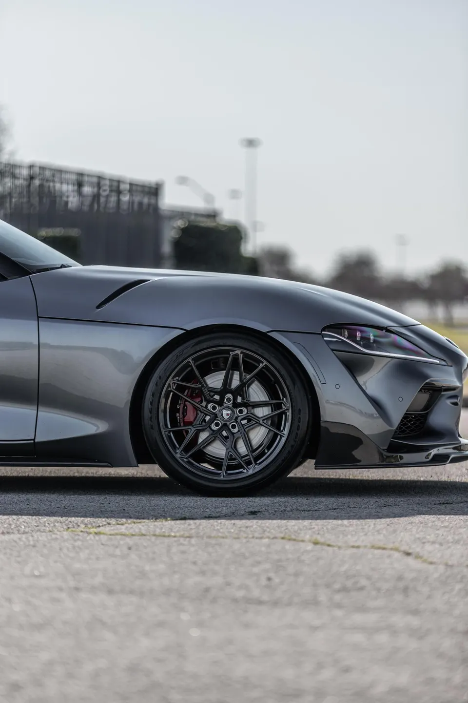 Close-up of a grey Toyota Supra's front wheel, custom black multi-spoke wheel, red brake caliper, and headlight.