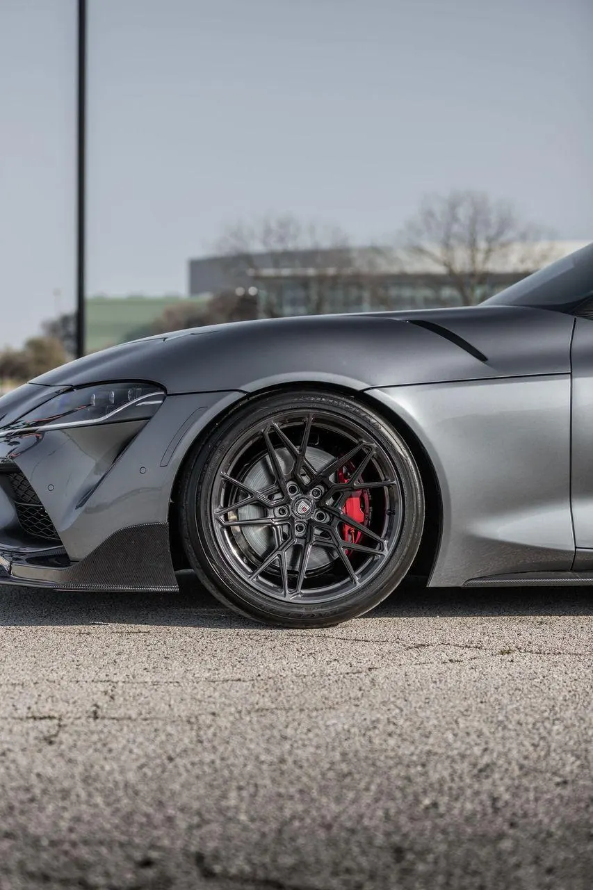 A close-up of a grey Toyota Supra's front wheel, showcasing custom dark multi-spoke wheels, red brake calipers, and carbon fiber aero.