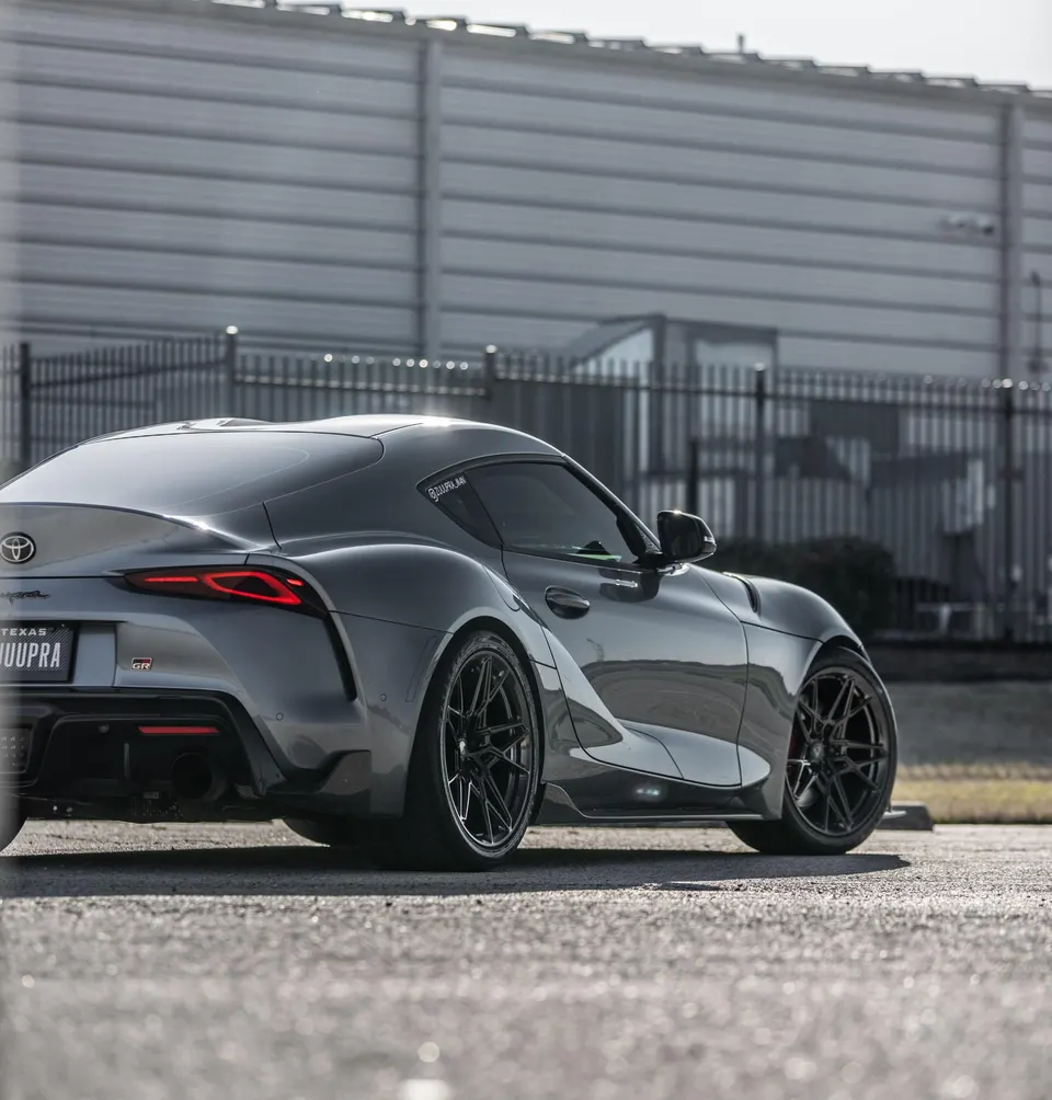 A grey Toyota Supra MK5 with aftermarket black wheels and a custom exhaust system parked in an industrial setting.