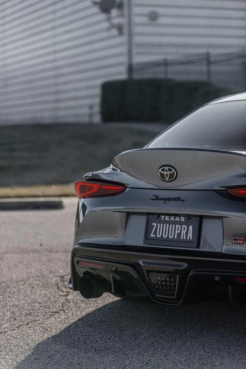 Rear view of a grey Toyota GR Supra with a carbon fiber diffuser, large exhaust tip, and custom Texas ZUUUPRA license plate.