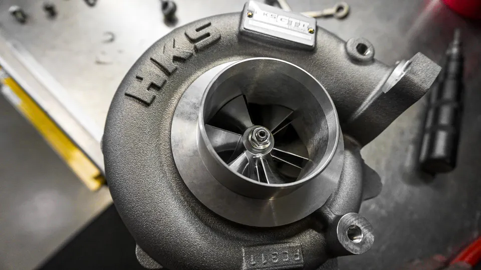 A close-up of an HKS turbocharger on a metallic surface, showcasing its compressor wheel and housing.
