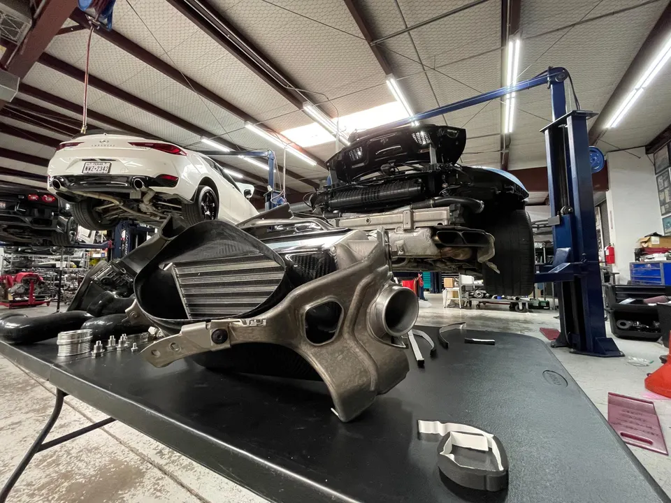 Intercooler and other performance parts on a table in a car shop with an Infiniti and Porsche on lifts.