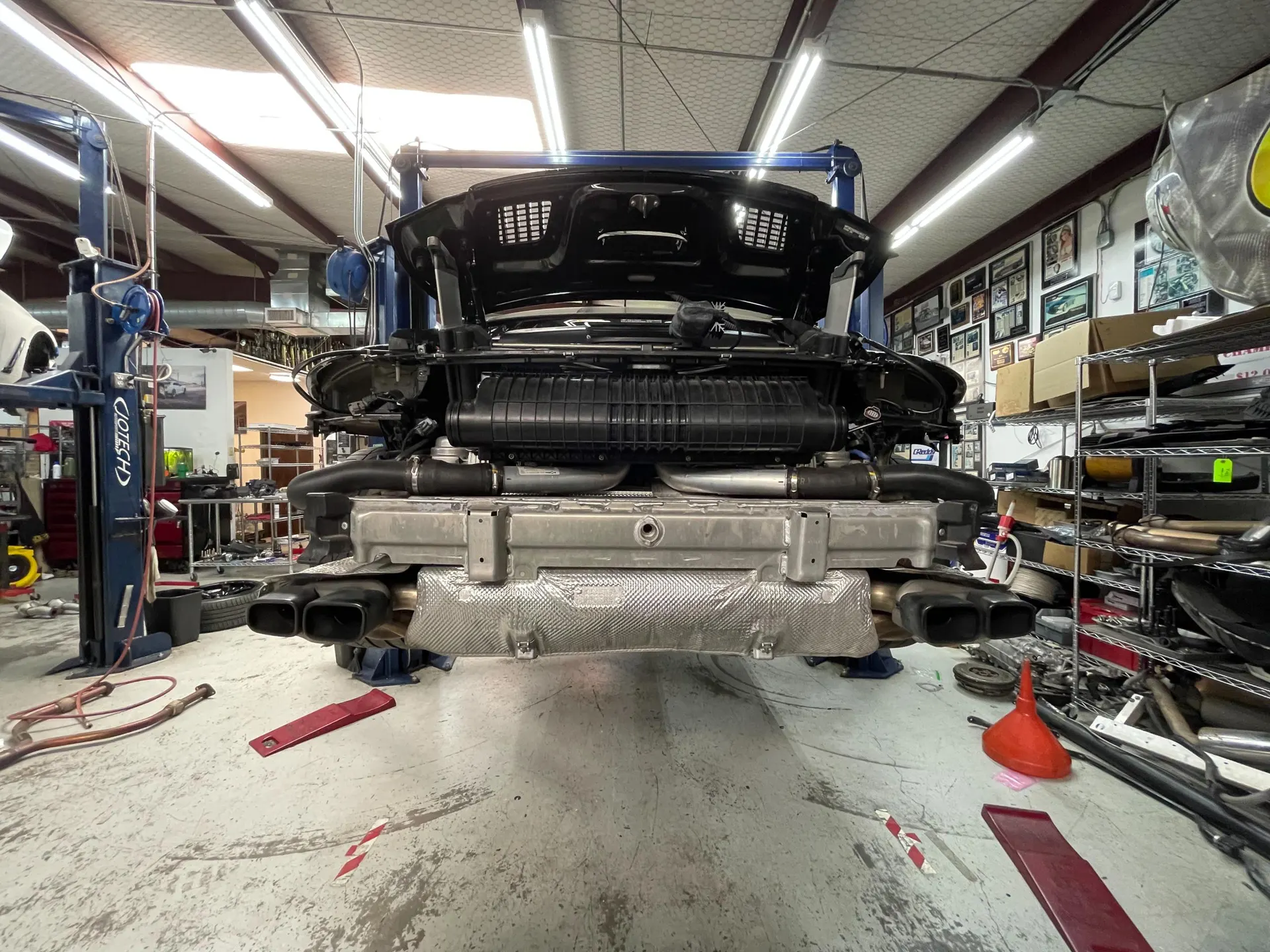Porsche 911 on a lift in a performance shop with the rear bumper removed, revealing the exhaust system and intercooler.