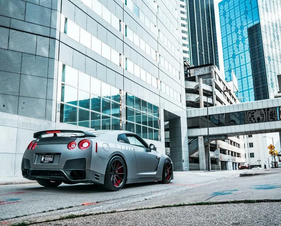 Matte grey Nissan GT-R with red wheels parked on a city street in front of modern buildings.