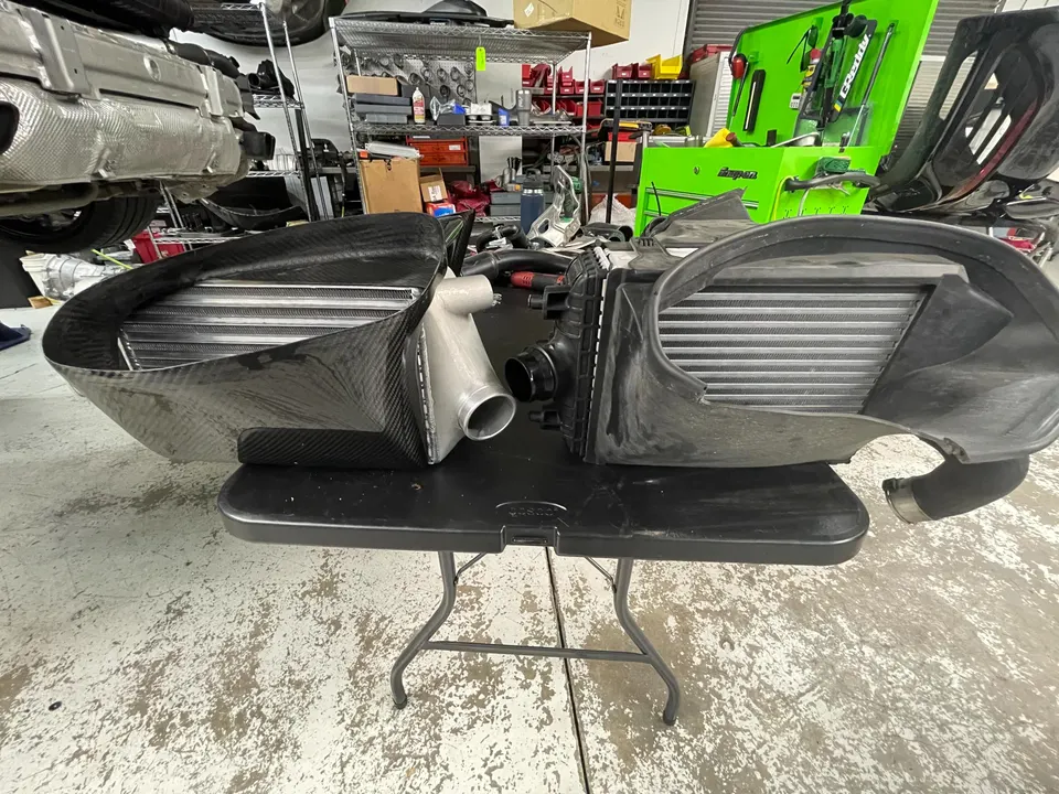 Two intercoolers, one carbon fiber aftermarket and one stock, displayed on a table in a performance shop.