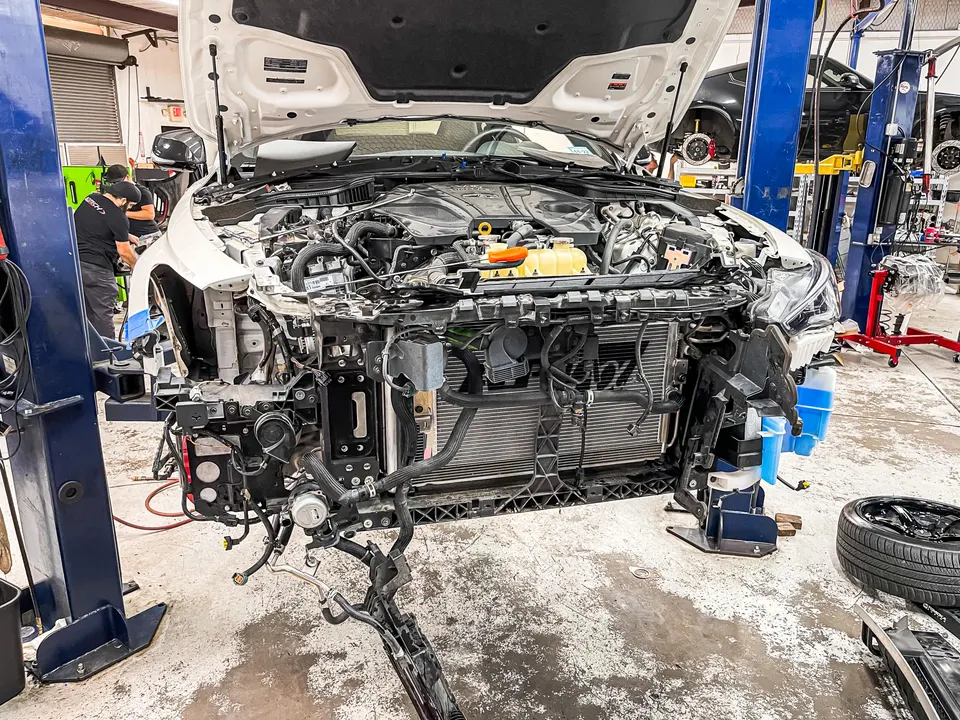 A white car with its front bumper removed and hood open, on a lift in a Jotech Motorsports shop, with mechanics working.