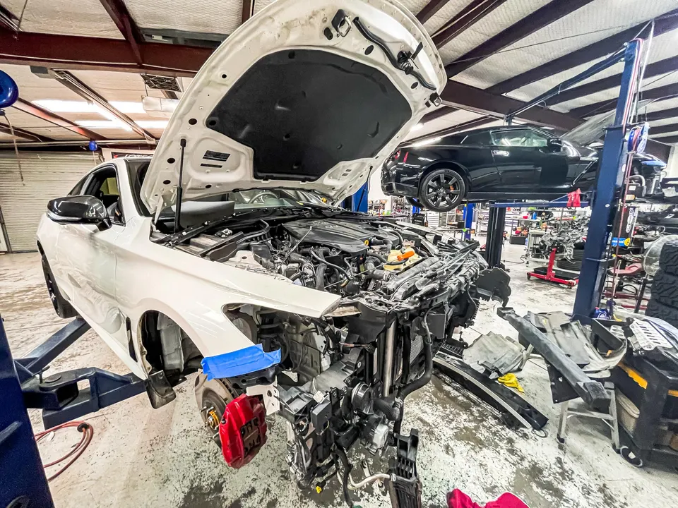 White car with its front end disassembled and hood open on a lift in a performance tuning shop, with a black car on another lift.