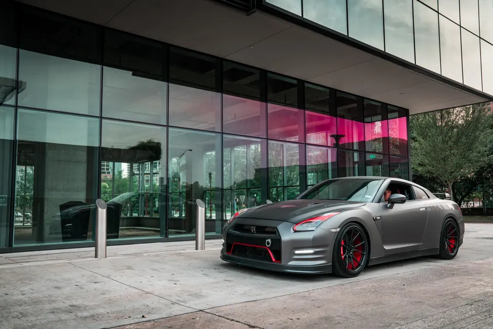 Grey Nissan GT-R with red accents and black wheels parked in front of a modern glass building in an urban setting.