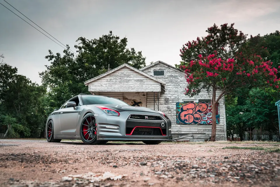 A grey Nissan GT-R with red accent wheels and front lip parked in front of an old, weathered house with graffiti.
