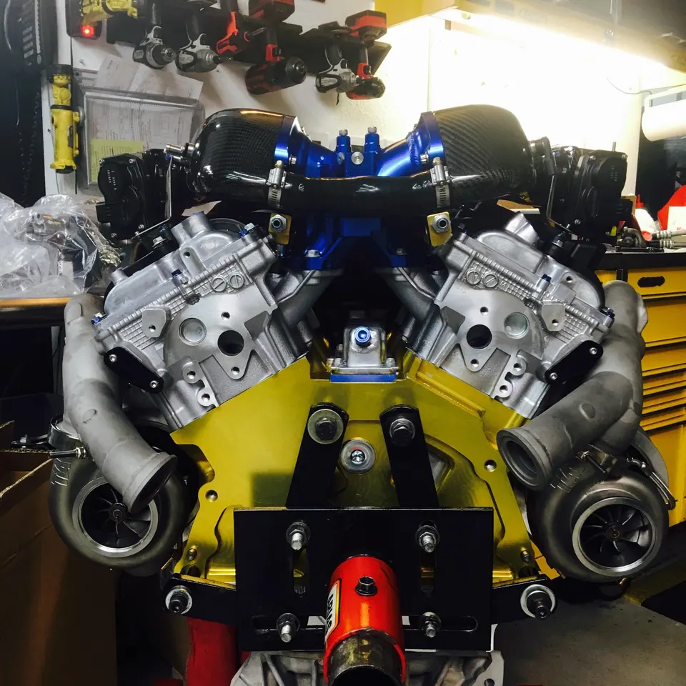 High-performance twin-turbo V-engine assembly with custom gold block, blue intake manifold, and carbon fiber intakes in a shop.