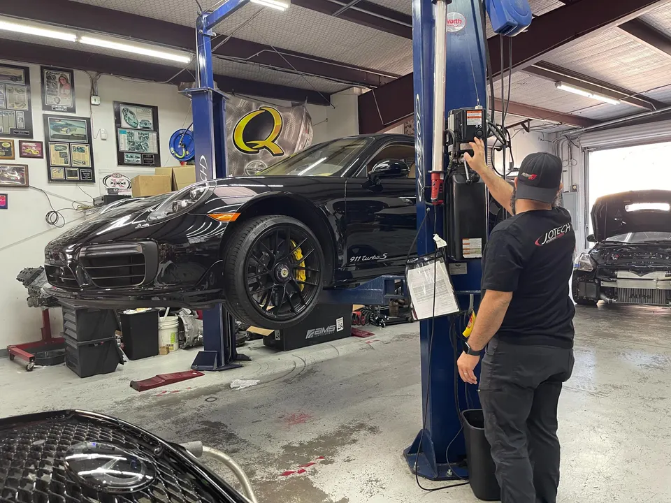 A black Porsche 911 Turbo S on a car lift in a Jotech Motorsports shop with a mechanic operating the lift.