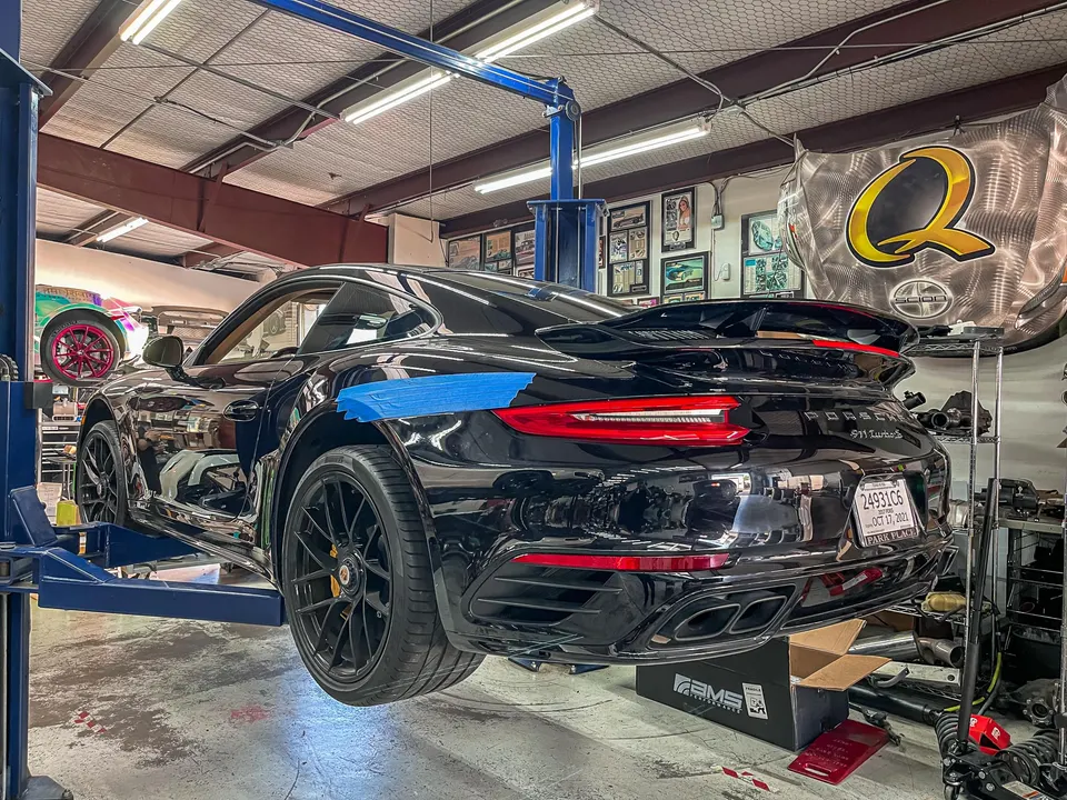 A black Porsche 911 Turbo S on a blue car lift in a performance tuning shop, with an AMS Performance box on the floor.