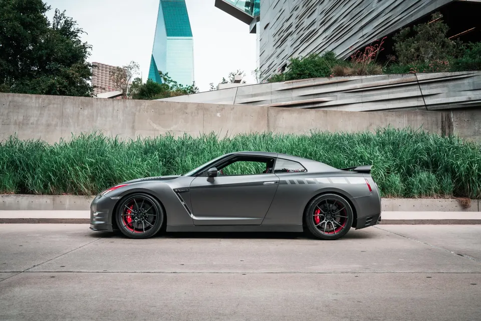 Grey Nissan GT-R R35 with aftermarket wheels and red calipers parked on a city street with modern architecture.