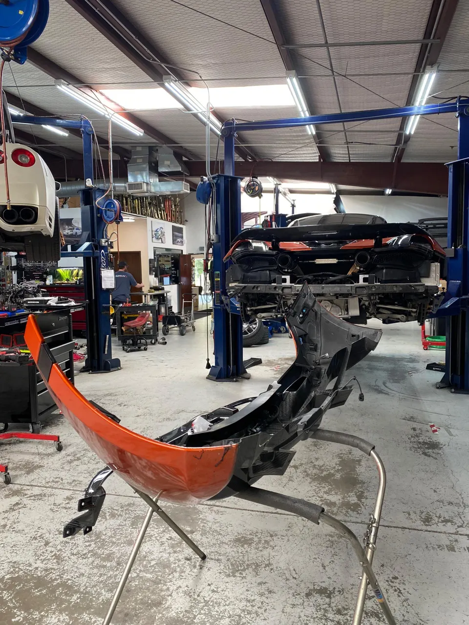 A McLaren supercar undergoing disassembly in a performance tuning shop, with its orange rear bumper on a stand.