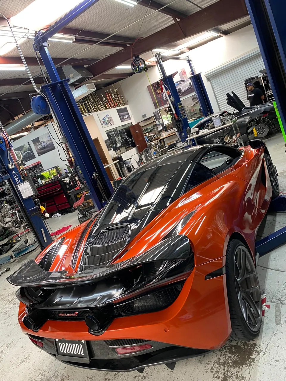 Orange McLaren 720S on a lift in a performance tuning shop, showcasing its carbon fiber rear wing and exhaust.