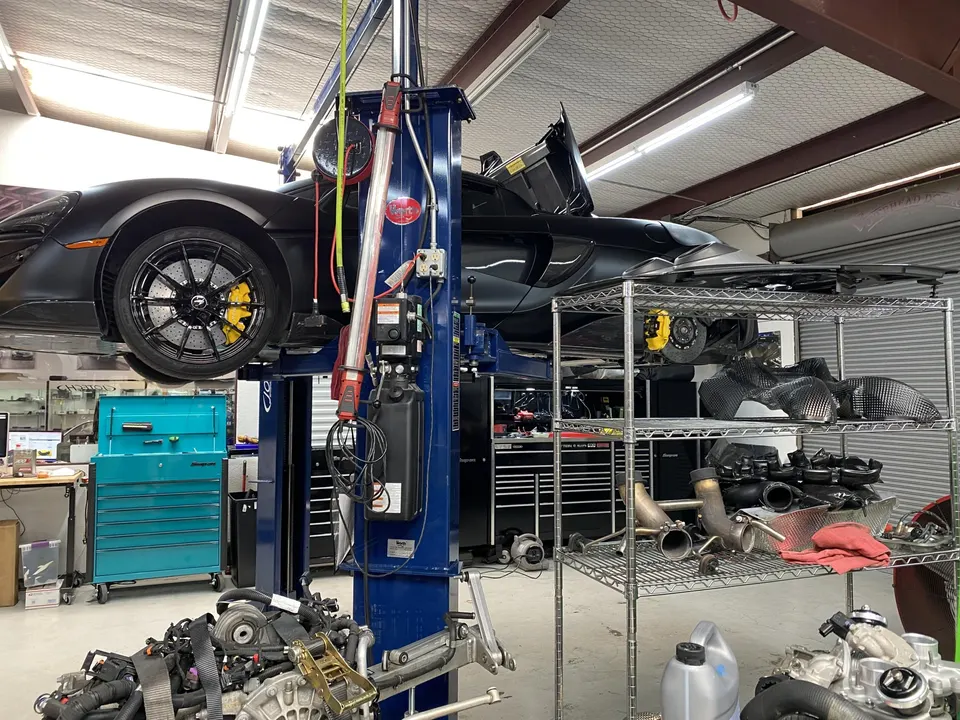 Black McLaren on a car lift in a performance tuning shop with an engine on the floor and various parts on shelves.