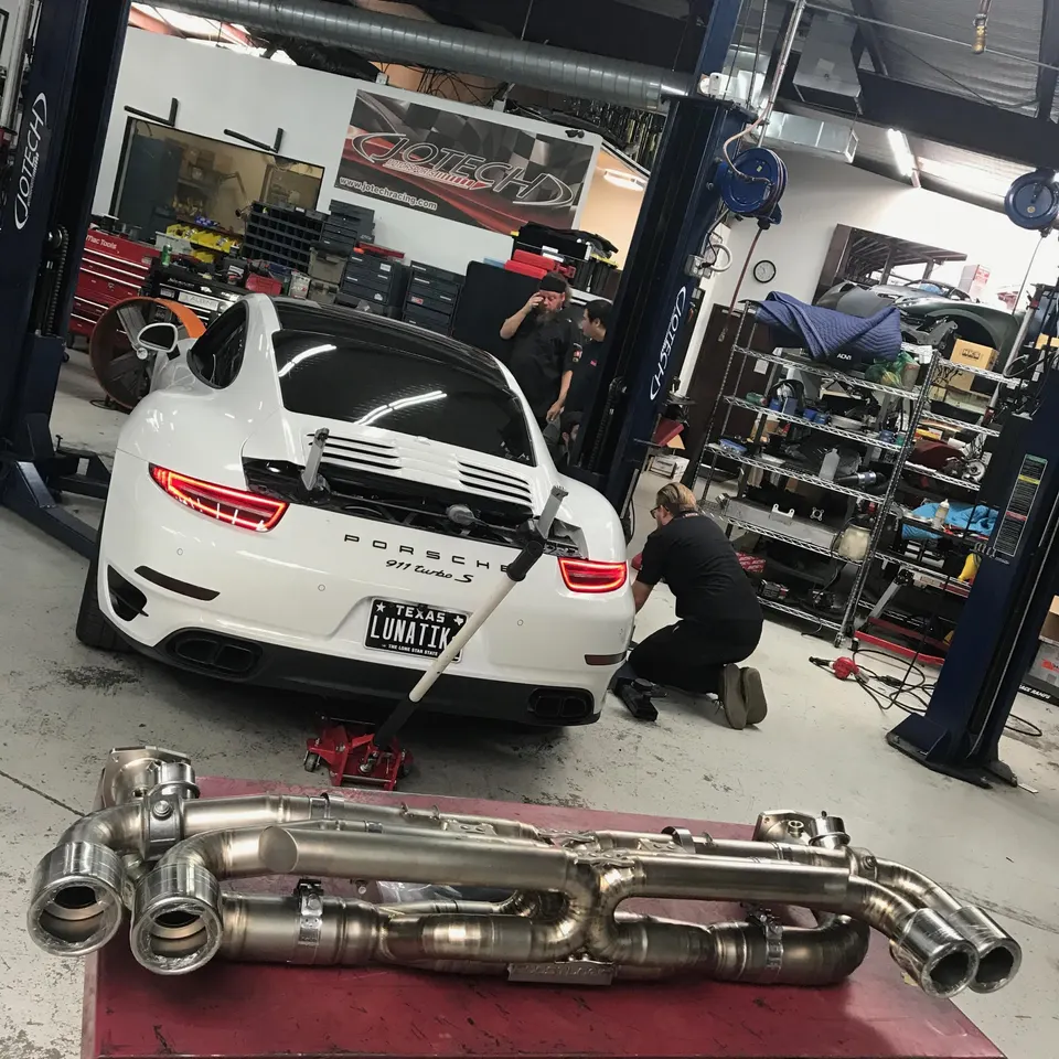White Porsche 911 Turbo S in a Jotech Racing shop with an aftermarket exhaust system on a table, being worked on by technicians.