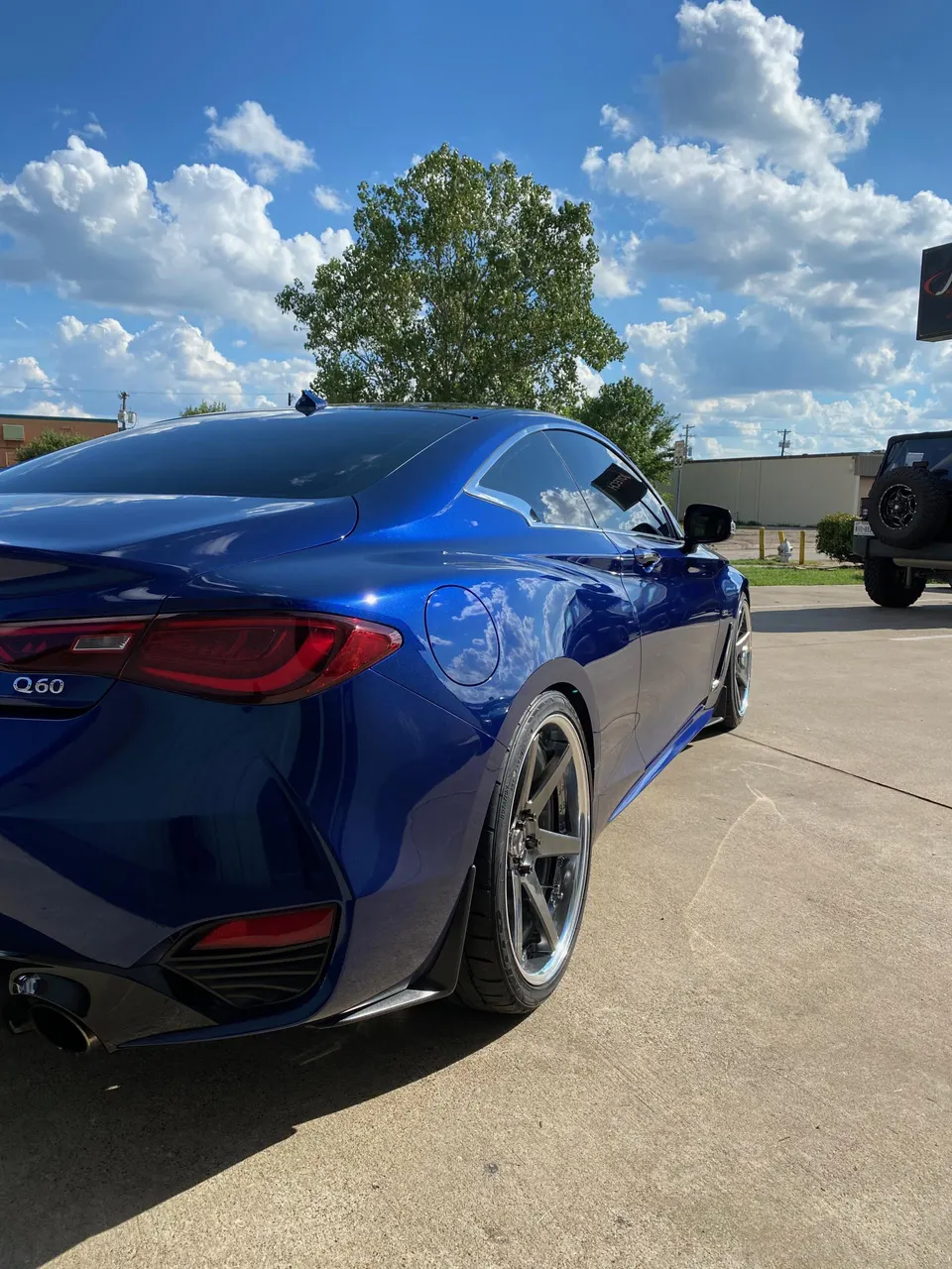 Blue Infiniti Q60 coupe with aftermarket wheels parked outdoors under a blue sky with clouds.