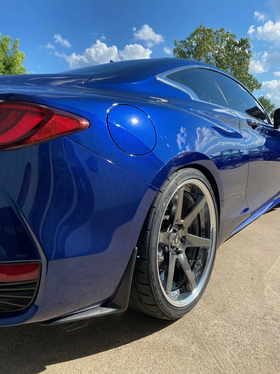 Blue Infiniti Q60 coupe with aftermarket multi-spoke wheels and performance tires, parked outdoors on a sunny day.