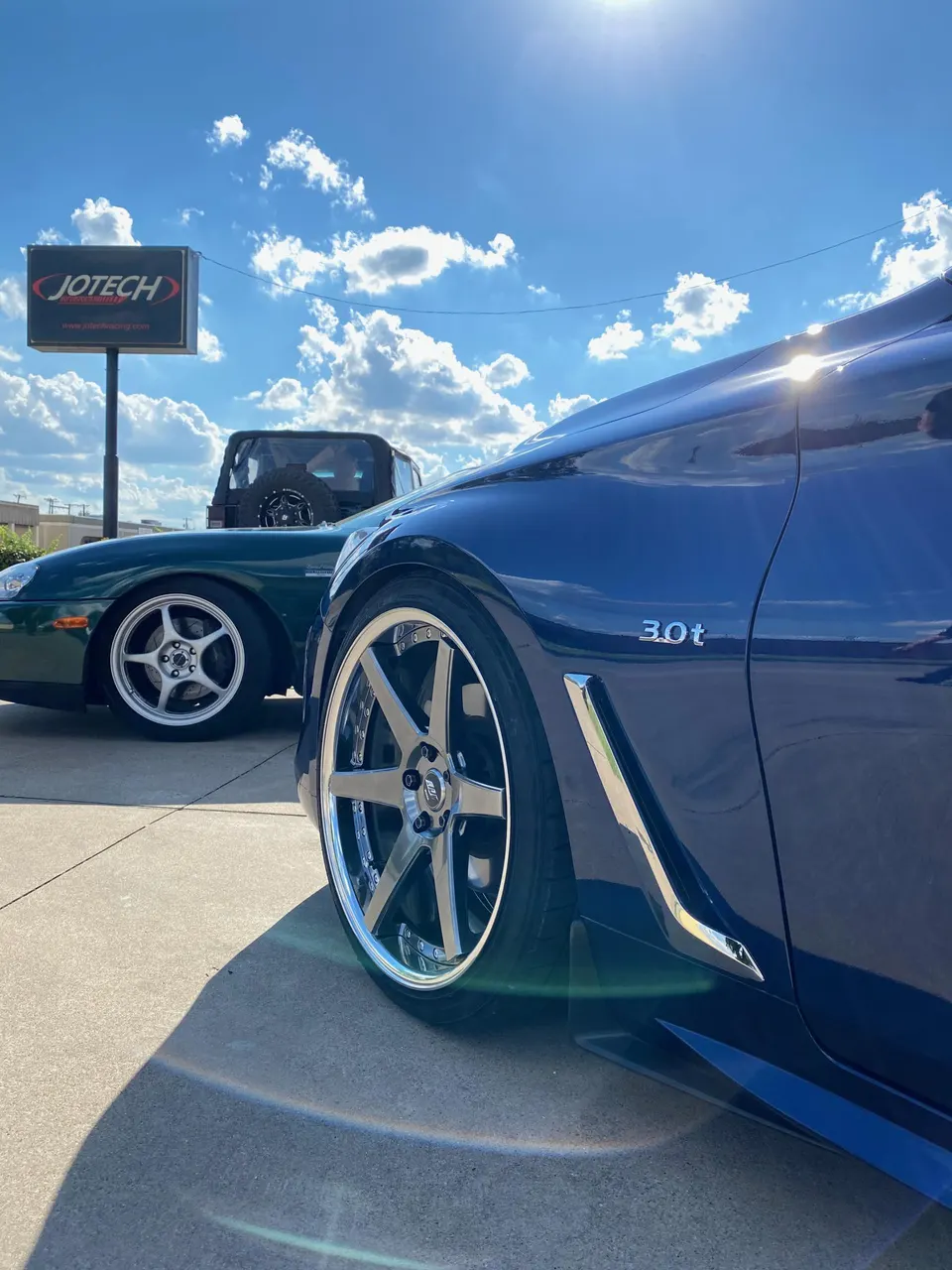 Blue car with custom chrome wheels parked outside Jotech Motorsports shop under a sunny sky, with a green car in the background.