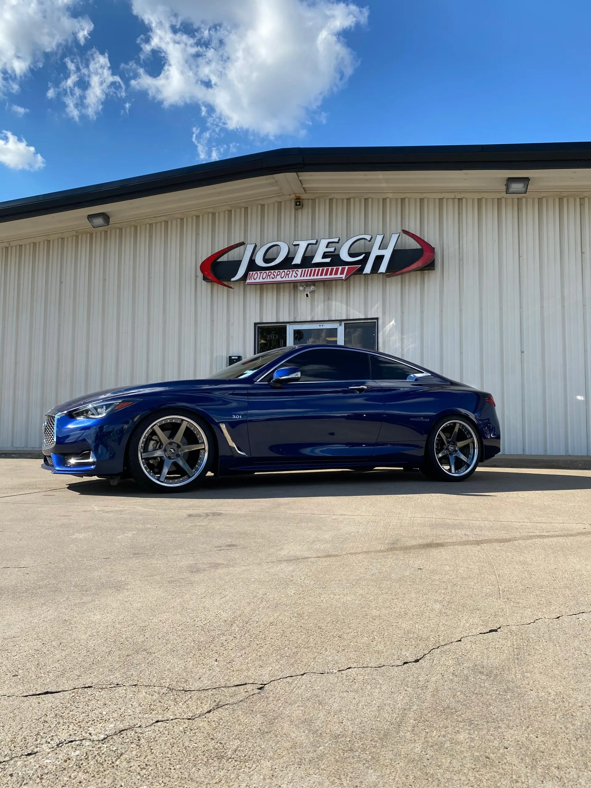 A blue Infiniti Q60 coupe with custom wheels parked in front of the Jotech Motorsports shop building.