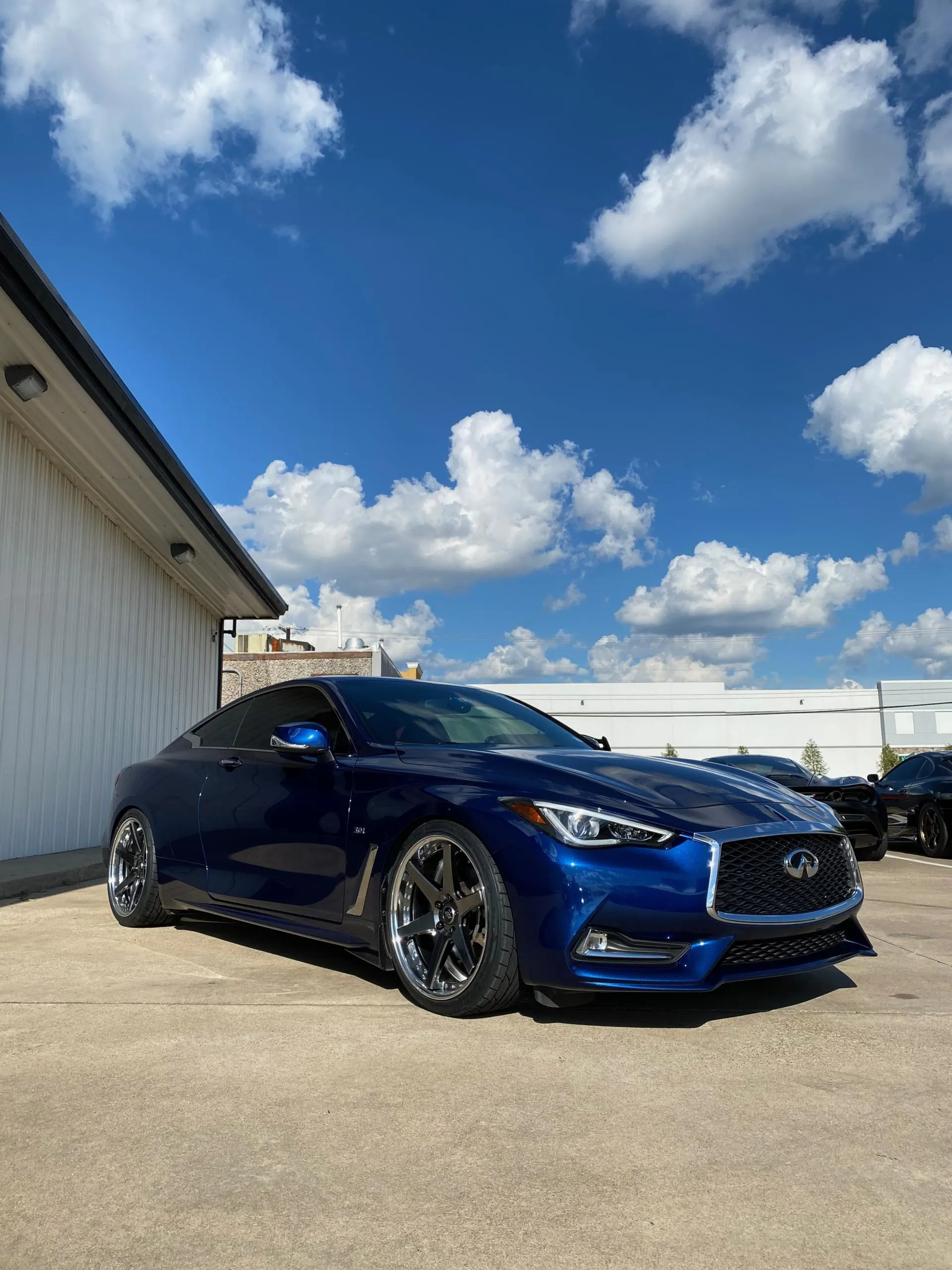 A blue Infiniti Q60 coupe with custom wheels parked outdoors under a blue sky.