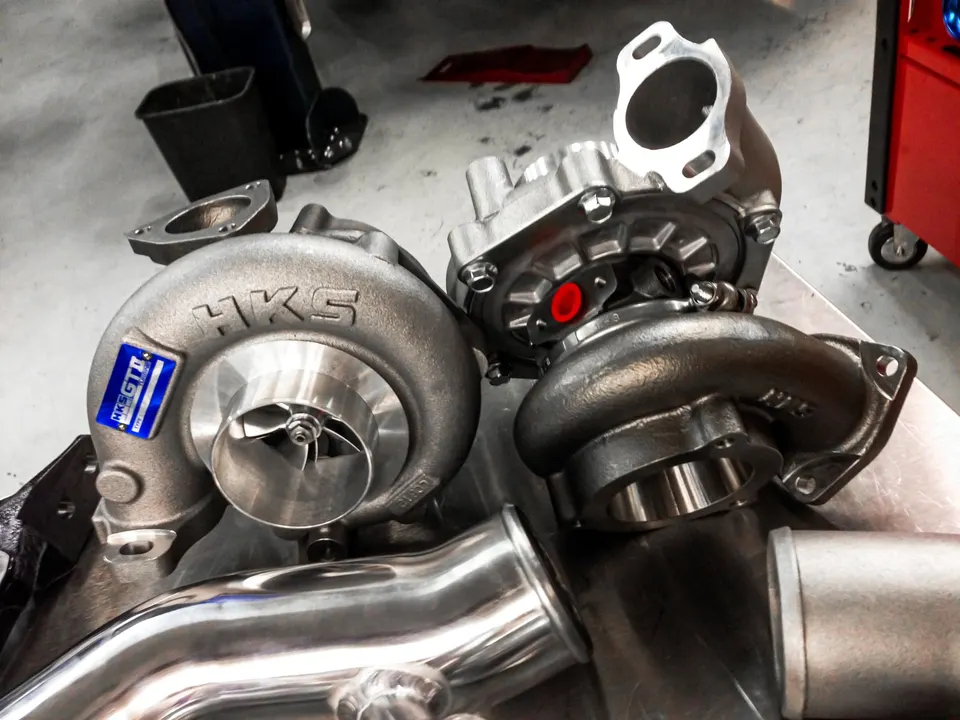 Two HKS turbochargers and polished exhaust piping on a workbench in a performance tuning shop.