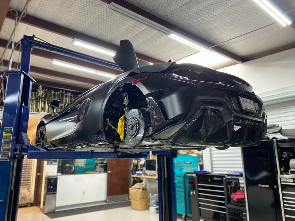 A black McLaren 720S on a car lift in a performance tuning shop, with the rear wheel removed exposing yellow brake calipers.