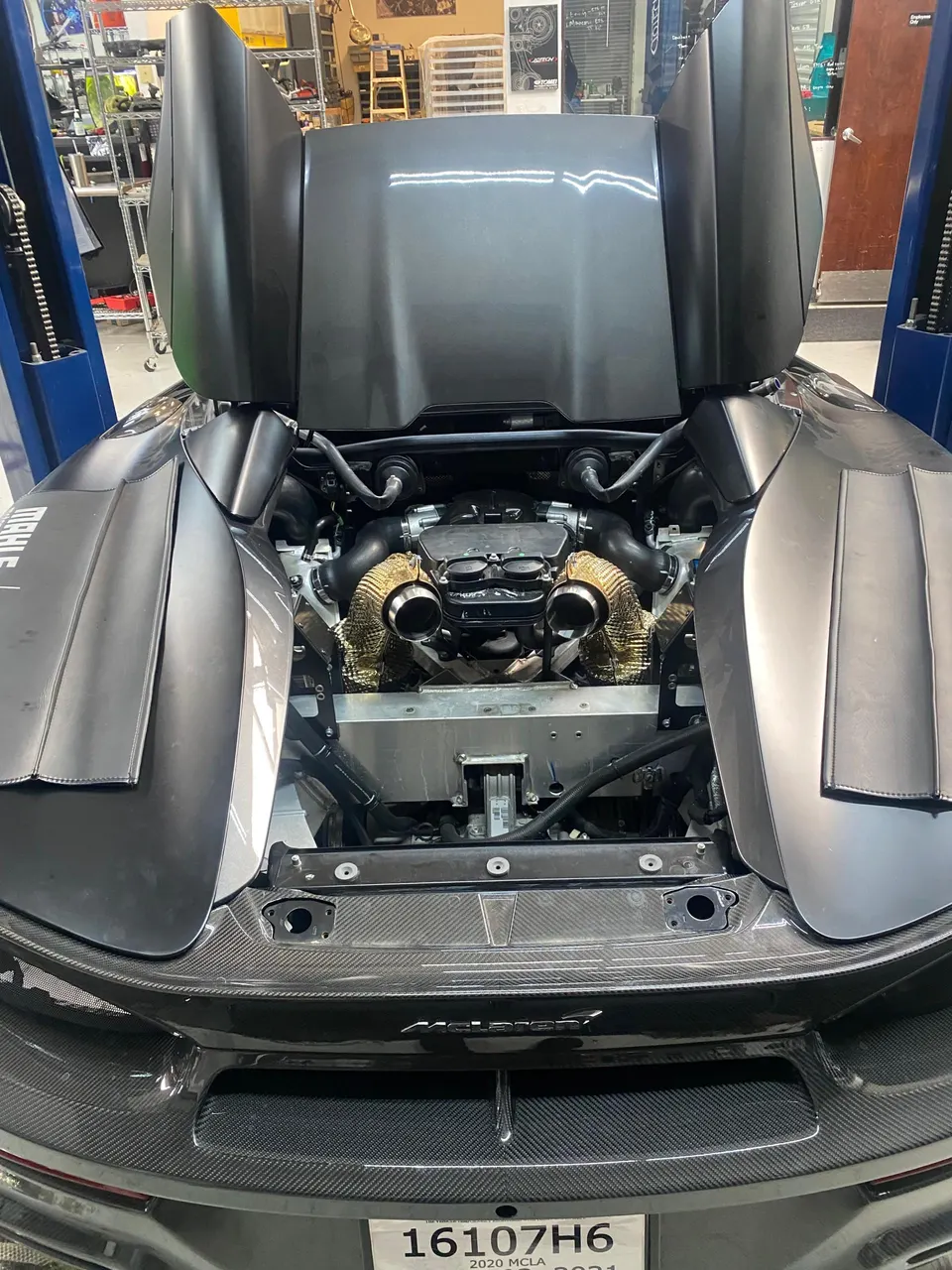 A McLaren supercar with its engine bay open, showcasing aftermarket turbochargers with gold heat shields in a performance shop.