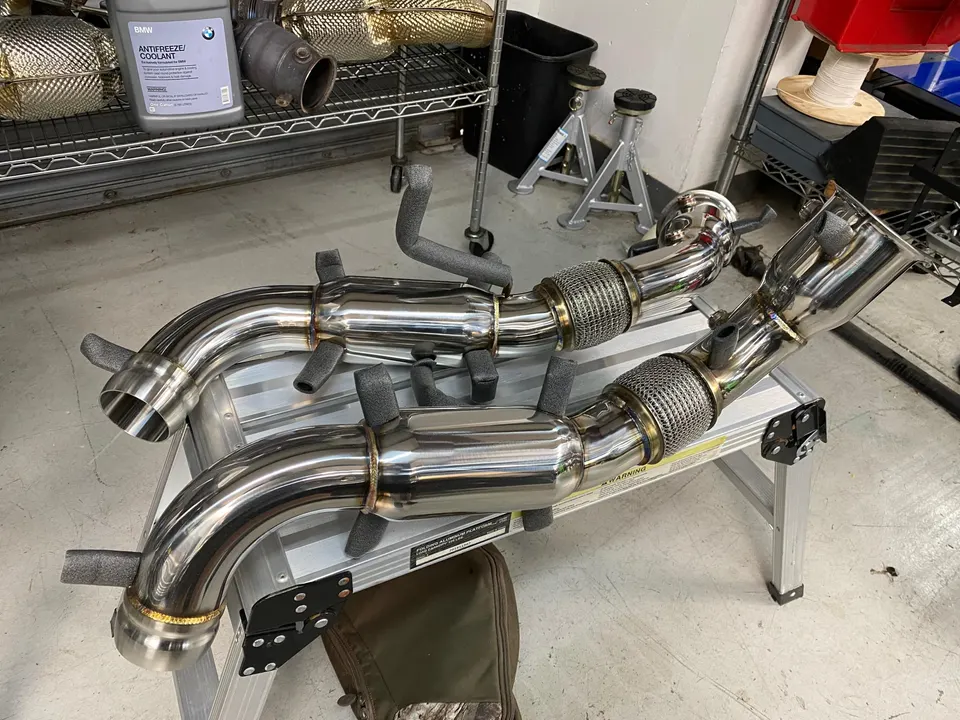 Two polished stainless steel performance exhaust downpipes with flex sections displayed in a shop environment.