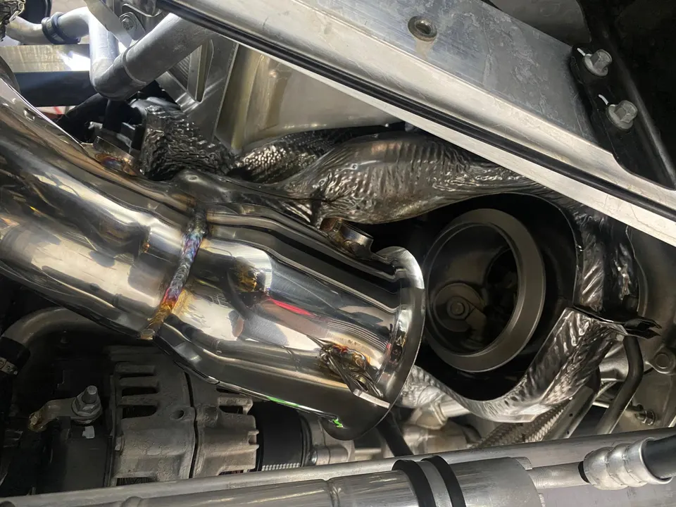Close-up of a custom polished downpipe with visible welds connected to a turbocharger in an engine bay.