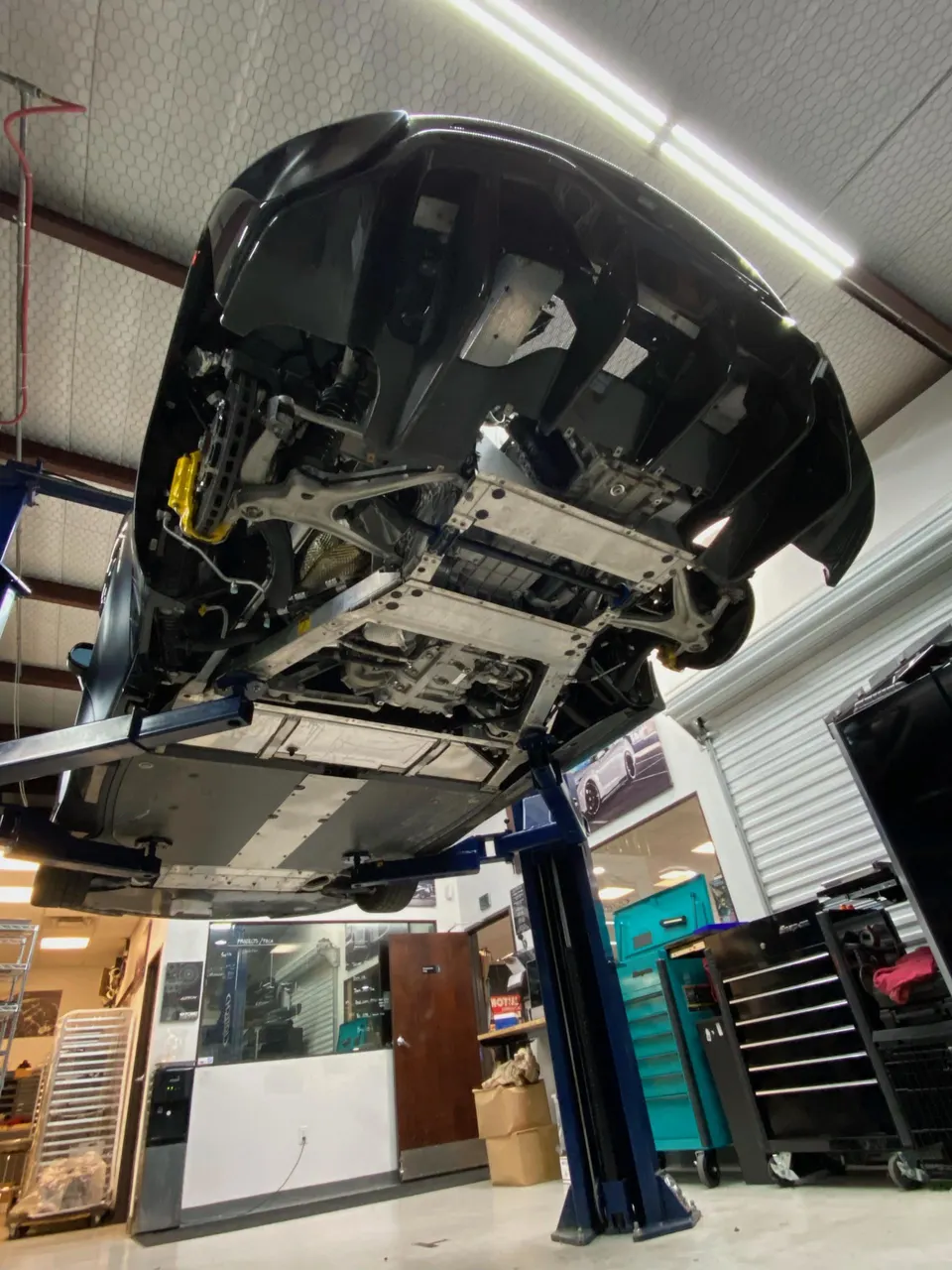 Underside view of a McLaren supercar on a lift in a performance tuning shop, showing suspension, yellow brake calipers, and rear diffuser.