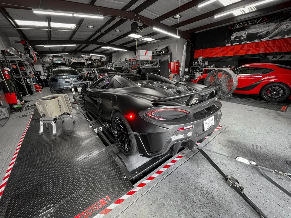 A matte black McLaren 570S convertible strapped to a dynamometer in a performance tuning shop with other cars in the background.