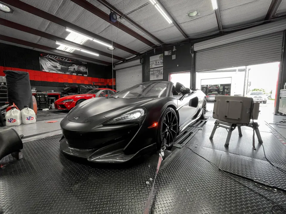 A matte black McLaren supercar strapped to a dynamometer in a performance tuning shop, with a red Toyota Supra in the background.