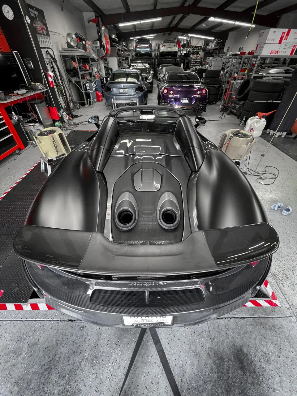Matte black McLaren 720S with a carbon fiber rear diffuser and dual exhaust tips in a performance tuning shop.